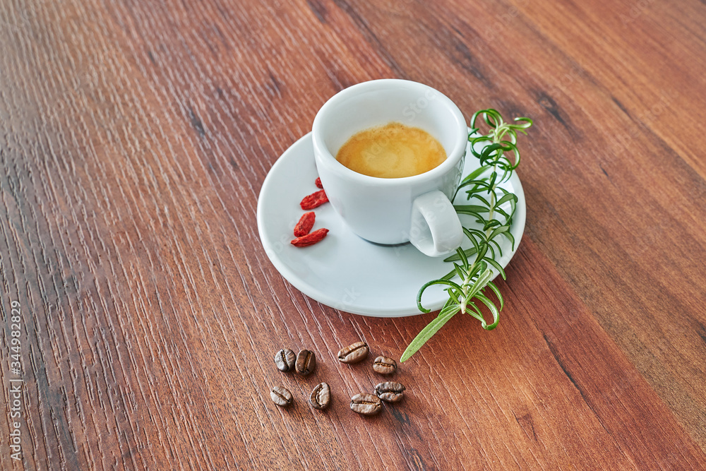 Italienischer Espresso in einer weißen Tasse mit Rosmarin, Goji Beeren und Kaffee Bohnen auf braunem Hintergrund. Italienischer Espresso in einer weißen Tasse mit Rosmarin, Goji Beeren und Kaffee Bohnen auf braunem Hintergrund.