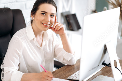 Portrait of a young attractive smiling business woman in a white shirt sitting at her workplace and working remotely at home