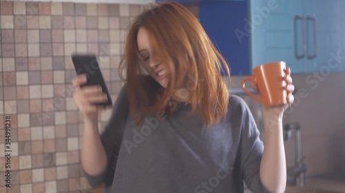 Girl with red hair in morning in kitchen listens to music, sings and dances. Young redhead woman dancing with a cup of coffee and a phone in her hands. Happy hipster millennial girl in t-shirt.