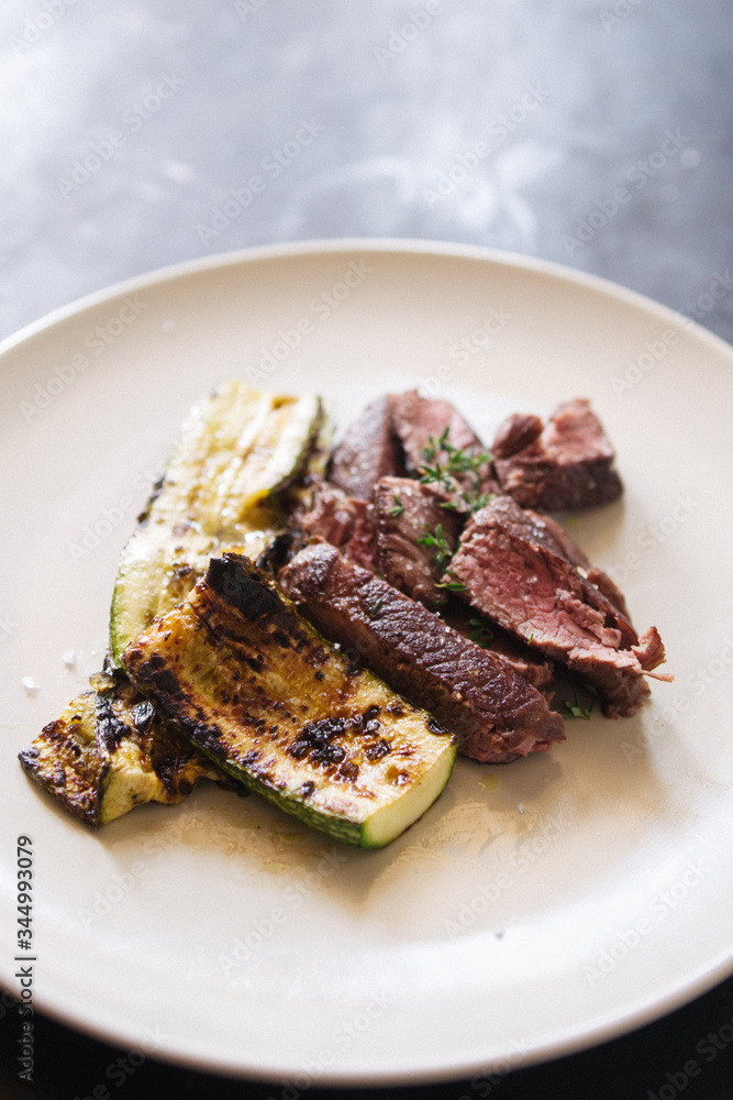 Meat fillet with grilled zucchini on a white dish with a white knife, top view over a black rustic board Meat fillet with grilled zucchini on a white dish with a white knife, top view over a black rustic board