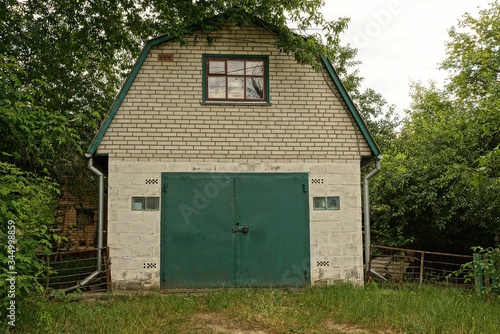 facade of a white brick garage with green metal gate and a window overgrown with green vegetation and grass on a rural street © butus