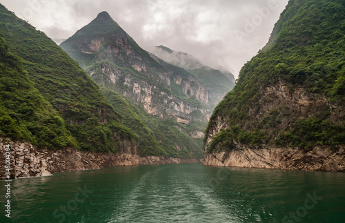 Wuchan, China - May 7, 2010: Dawu or Misty Gorge on Daning River. View into canyon between tall green covered mountains with some cliff sides around emerald green water under cloudscape. © Klodien