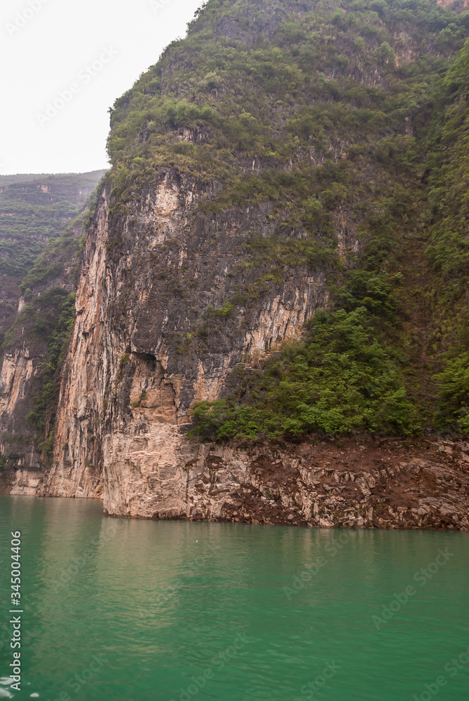 Wuchan, China - May 7, 2010: Dawu or Misty Gorge on Daning River. Brown rocky cliffs descends as wall into emerald green water along canyon. Some green foliage and black covered stones.