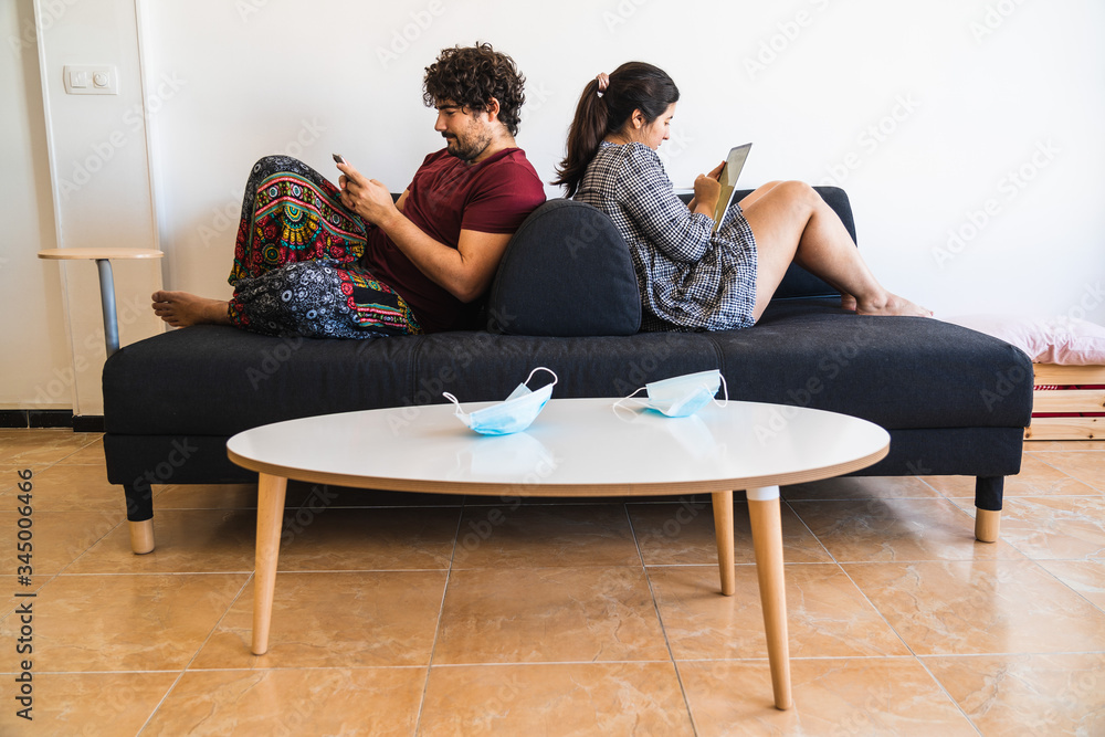 Stockfoto New normal situation after the coronavirus covid 19 sanitary  crisis, a spanish couple rest on a couch reading and using a smartphone to  chat while their surgical masks rest at the