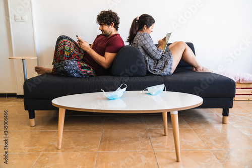 New normal situation after the coronavirus covid 19 sanitary crisis, a spanish couple rest on a couch reading and using a smartphone to chat while their surgical masks rest at the living room table