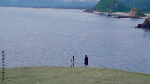 Panning shot of girl and boy walking on field by sea against sky, idyllic view of mountains during sunset - Lombok, Bali photo