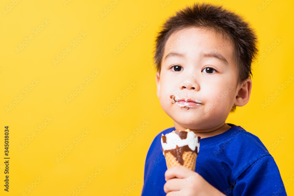 Happy portrait Asian child or kid cute little boy attractive laugh smile playing holds and eating sweet chocolate ice cream waffle cone, studio shot isolated on yellow background, summer concept Happy portrait Asian child or kid cute little boy attractive laugh smile playing holds and eating sweet chocolate ice cream waffle cone, studio shot isolated on yellow background, summer concept