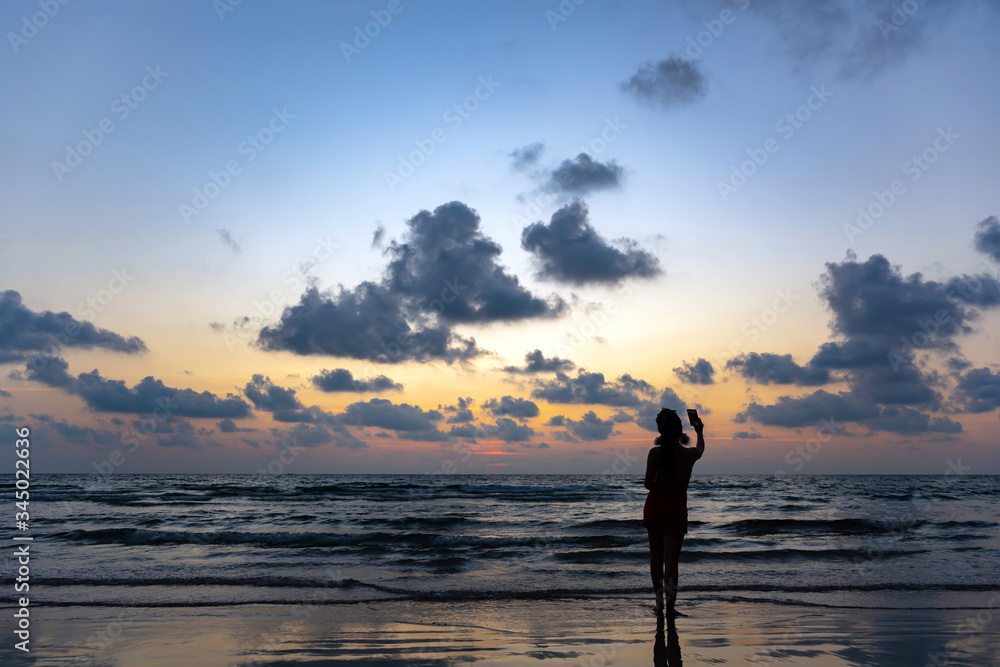The silhouette of a single woman using a selfie to take a picture of herself By the sea at sunset, beaches and beautiful sky at Ko Kood, Thailand
