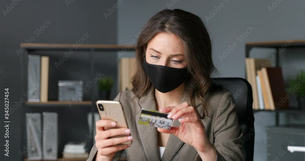 Young Caucasian female sitting at table in office with credit card in hand and shopping online on smartphone. Pretty businesswoman in mask buying in internet on phone and paying. Shopper on quarantine Young Caucasian female sitting at table in office with credit card in hand and shopping online on smartphone. Pretty businesswoman in mask buying in internet on phone and paying. Shopper on quarantine