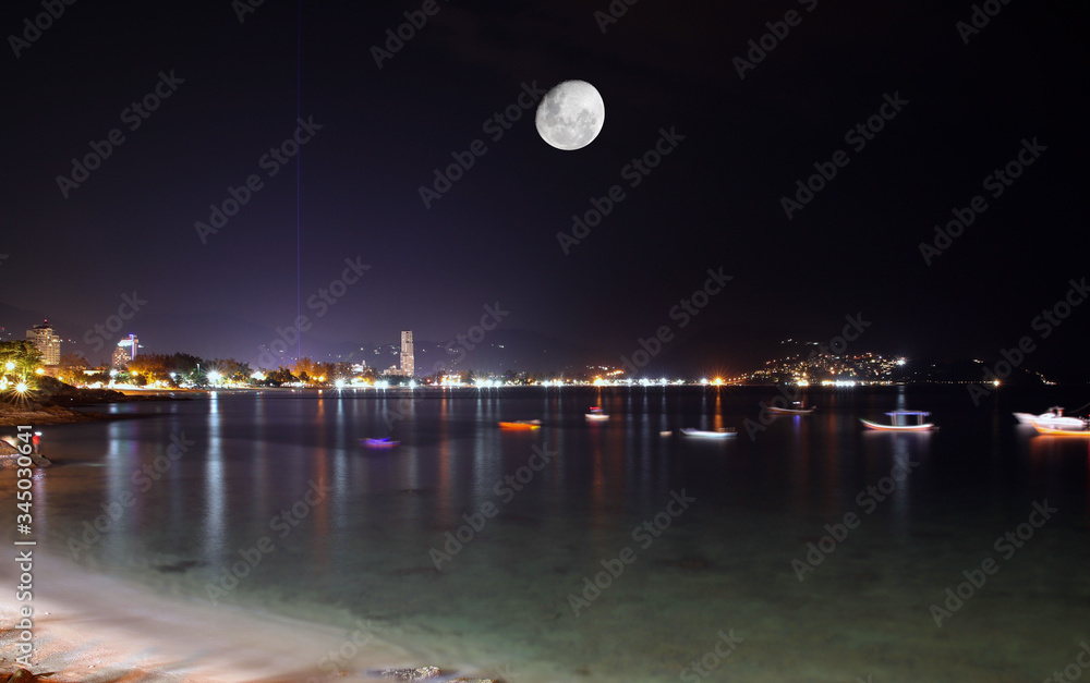 Patong Phuket Thailand night time long expose photograph with the night skyline illuminated by the city lights and the moon over the mountains