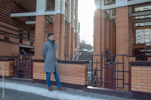 stylish guy in grey overcoat in leather boots poses holding hands behind back standing against modern building. Concept fashion city environment