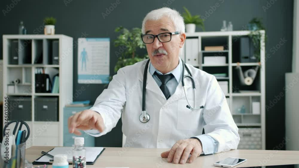 Portrait of joyful doctor senior man in uniform talking waving hand showing bottles with pills in hospital office during online call with patient. Medicine, people and consultation concept. Portrait of joyful doctor senior man in uniform talking waving hand showing bottles with pills in hospital office during online call with patient. Medicine, people and consultation concept.