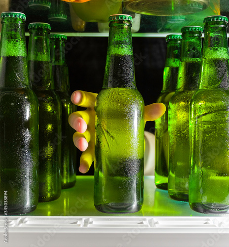 The hand reaches for the beer bottle. Green bottles of beer and drops of condensation are on a shelf in the refrigerator.