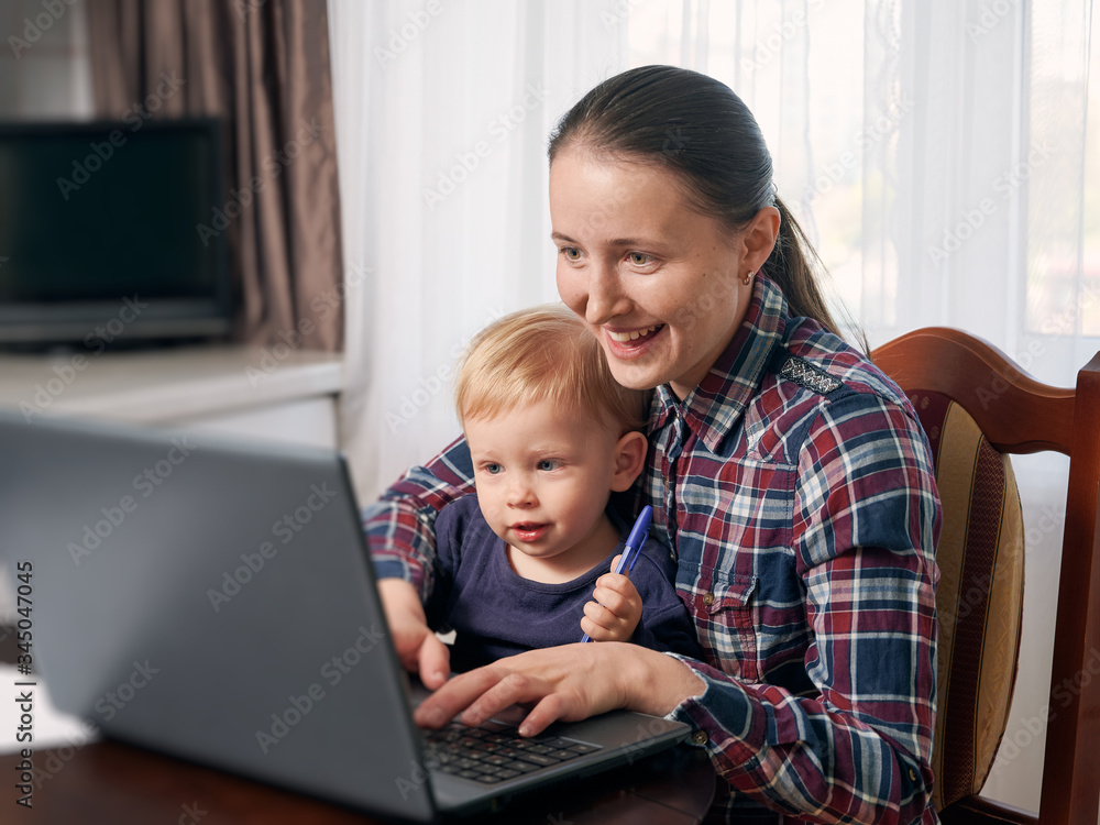 Happy mother working remotely on laptop while taking care of her little son. Young woman trying to freelance by the table with toddler on her laps. Quarantine isolation concept Happy mother working remotely on laptop while taking care of her little son. Young woman trying to freelance by the table with toddler on her laps. Quarantine isolation concept