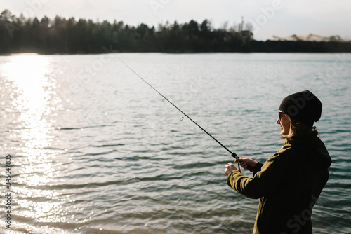Woman catching a fish, pulling rod while fishing. Girl fishing from beach lake or pond with text space.Fisherman with rod, spinning reel on the river bank. Sunset. Fishing for pike, perch, carp.