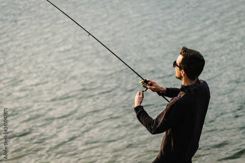 Man catching fish, pulling rod while fishing from lake or pond. Fisherman with rod, spinning reel on river bank. Sunrise. Fishing for pike, perch, carp on beach lake or pond. Background wild nature.
