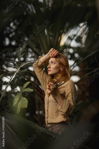 A beautiful girl with natural make-up and red hair stands in the jungle among exotic plants with snake.