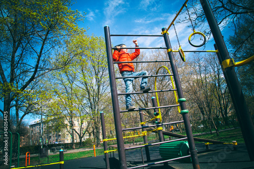 A boy in a red jacket is engaged in a sports field in the Park in the spring, climbing up the stairs. Swedish wall. Trips, sports classes for children, physical education, games in the fresh air.