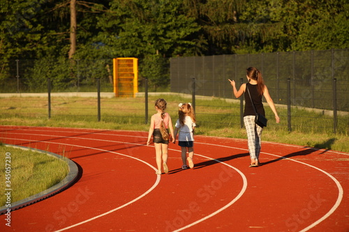 A young mother and her daughters are running along the treadmill of a stadium around a football field. Artificial cover of the sports complex. Parenting in healthy activity. Prevention of seasonal