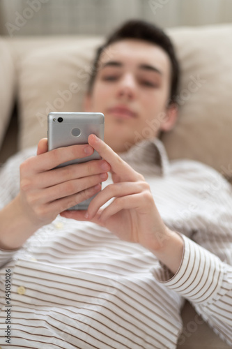 Teenager young man lying on sofa at home, holding and using smart phone