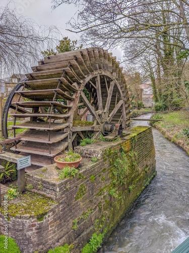 Old wooden water wheel along stream in rural village. Moss covers stone support. Sign informs that fishing is prohibited nearby. 