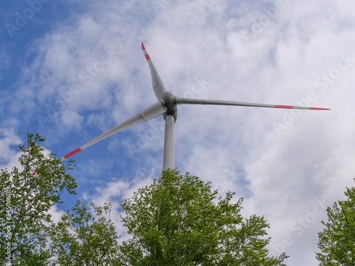 Windrad im Wald mit Blickpunkt nach oben nah am Windrad bei blauem Himmel mit weißen Wolken und hellgrünen Bäumen im Vordergrund