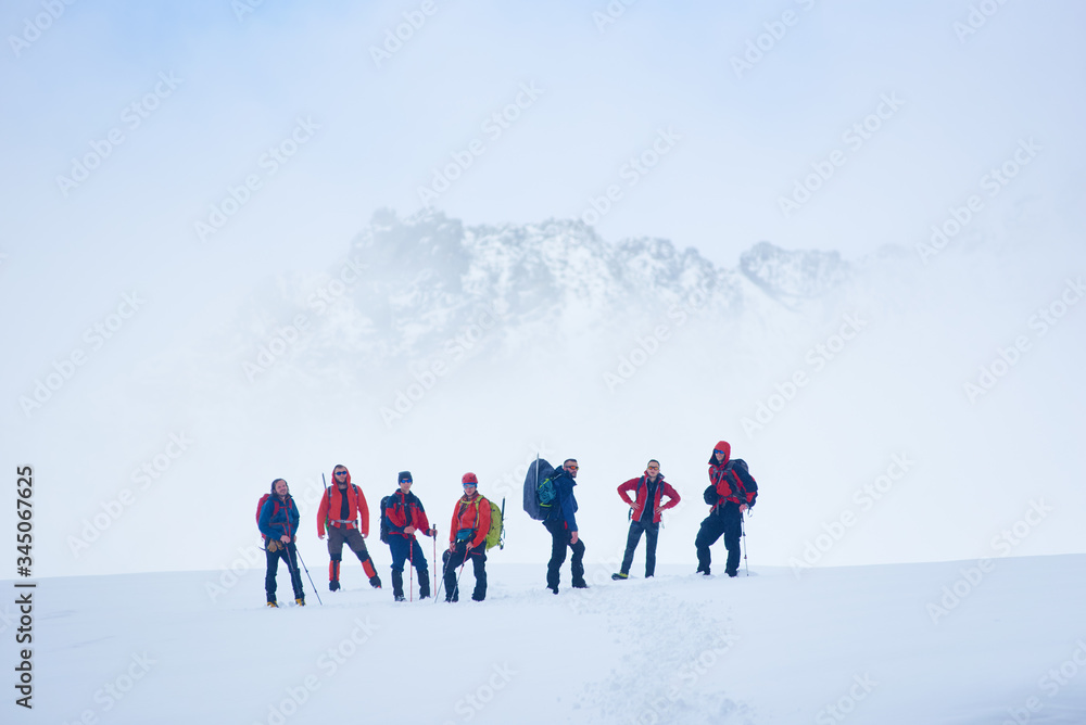 Hikers team standing on hillside valley covered with snow. Male alpinists and trekking sticks enjoying the view of winter mountains while taking rest break during hike, looking to the camera