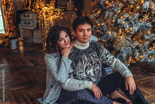 Couple of lovers married people on cozy sweaters hugging sitting under a Christmas tree in a house with a beautiful interior and a large window © Valeriia