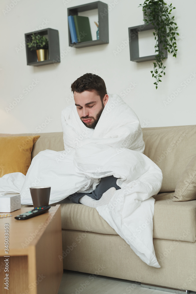 Sick guy wrapped into blanket sitting on couch in front of table with medicine
