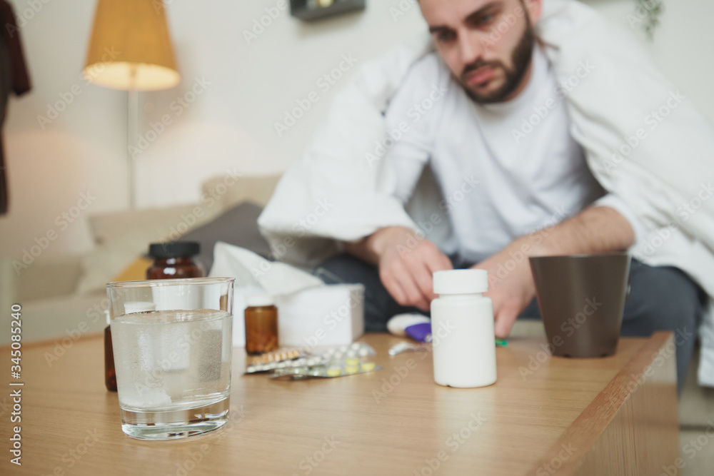 Sad and sick young man wrapped in blanket sitting in front of table with pills Sad and sick young man wrapped in blanket sitting in front of table with pills