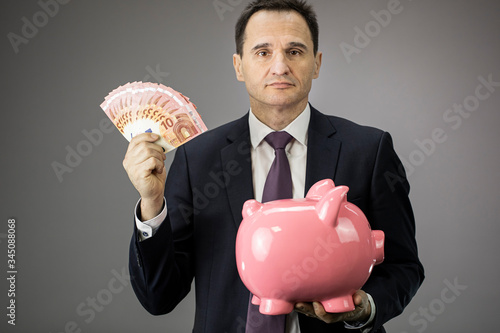 Confident successful businessman holds piggy band and money banknotes europen cash in hands, attractive middle aged man in formal clothes looking relaxed at camera. Income, savings, investment concept
