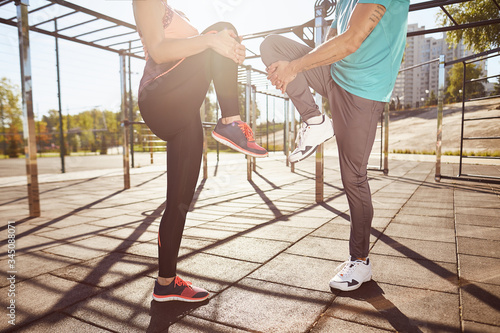 Stretching exercises. Cropped photo of man and woman in sportswear warming up together while standing at the stadium. They are stretching legs. Active couple doing sport outdoors