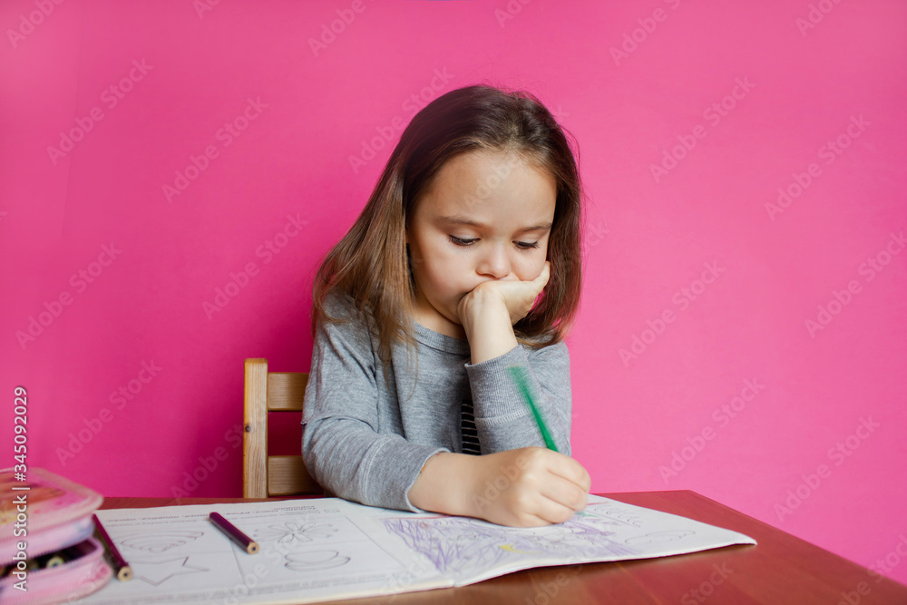 A cute little girl is sitting at a table and doing her homework. Pink background. Distance learning at home A cute little girl is sitting at a table and doing her homework. Pink background. Distance learning at home