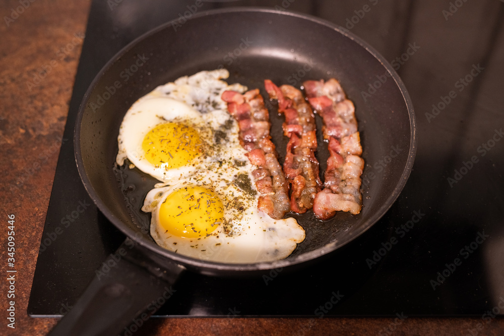 Fototapeta premium Frying pan with fried eggs and bacon on electric stove in process of cooking