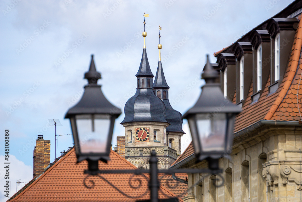 Kirchturm mit schwarzem Schieferdach und rot goldene Uhr über den Häuserdächern.Bayreuth. Kirche der Stadt Bayreuth zwischen zwei Laternen. Stadtkirche, Bayern, Deutschland. Klassische Architektur. Kirchturm mit schwarzem Schieferdach und rot goldene Uhr über den Häuserdächern.Bayreuth. Kirche der Stadt Bayreuth zwischen zwei Laternen. Stadtkirche, Bayern, Deutschland. Klassische Architektur.