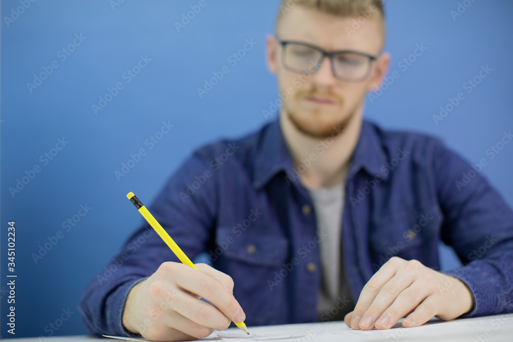 Young handsome architect at work, yellow pencil in hand in focus, man on blue background drawing construction plan, working on project from home office. Concept of architects, engineer holding pencil Young handsome architect at work, yellow pencil in hand in focus, man on blue background drawing construction plan, working on project from home office. Concept of architects, engineer holding pencil