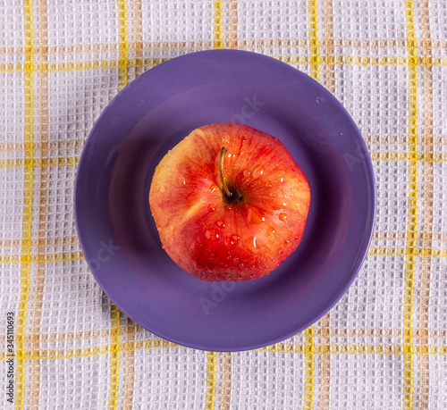 A red Apple on a purple plate on the table. Drops of water glisten on the Apple.
