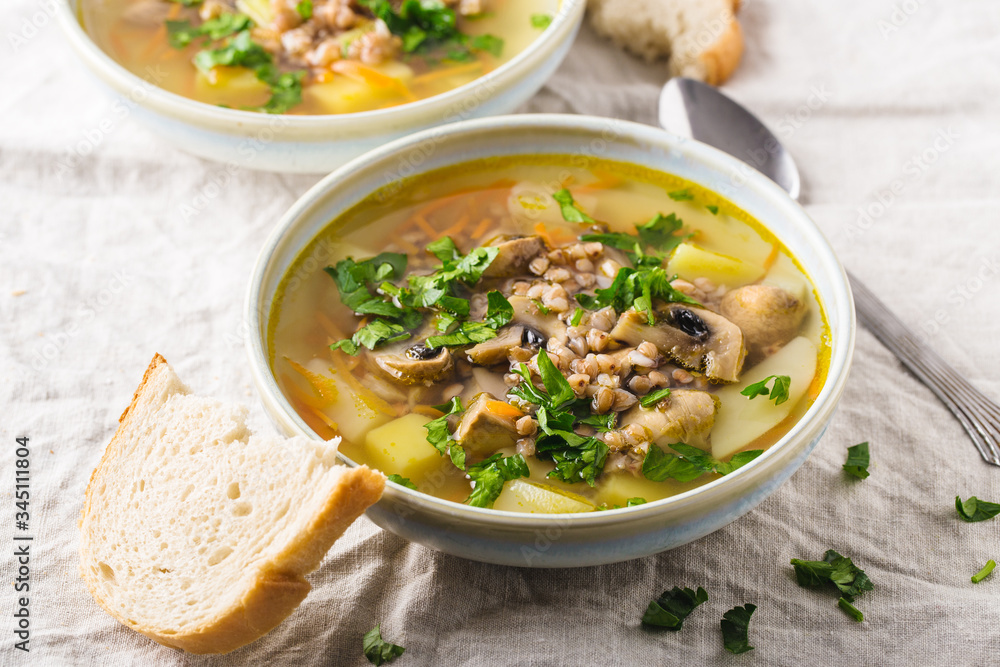 Soup of mushrooms, buckwheat, potatoes and carrots with herbs in bowls with slice of white bread on the side, light background, horizontal format. Top view