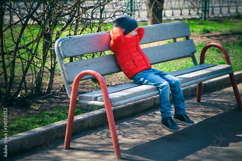 A boy in a red jacket sits on a Park bench in the spring, resting after outdoor games. He put his hands behind his head and swung his legs over the side. Walking with a child, playing in the fresh air