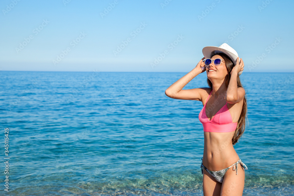 Beach vacation. Hot beautiful woman in sun hat and bikini standing with her arms raised to her head enjoying looking view of beach ocean on hot summer day.
