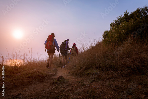 Girl with red backpack and friend is climbing, Two walkers are walking through beautiful meadows and bright sky.