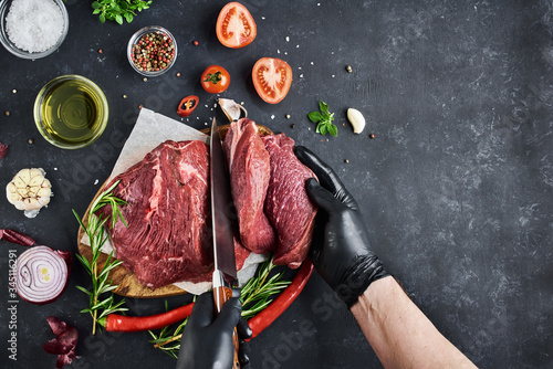 The butcher cuts portions of steaks from a large piece of fresh meat tenderloin, which lies on parchment on a dark background. On the table-spices, vegetables and herbs. Top view, free space for text.