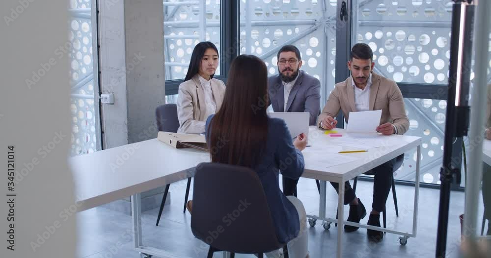 Mixed-races young employees having meeting with female boss while sitting at table in office. Multiethnic white-collars workers of firm discussing business and project with company head. Rear. Mixed-races young employees having meeting with female boss while sitting at table in office. Multiethnic white-collars workers of firm discussing business and project with company head. Rear.