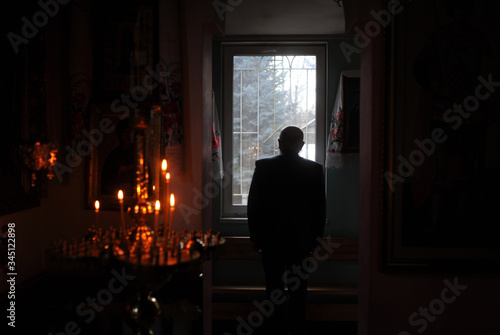 Silhouette of a priest standing before a bright window in the dark room with candles.