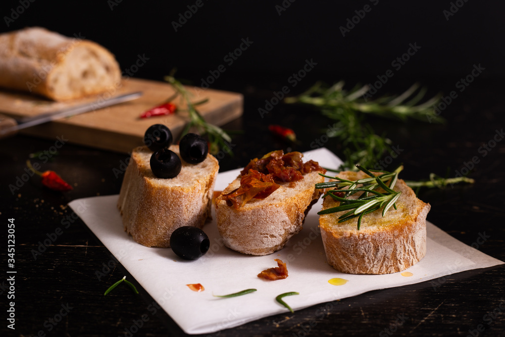 Sliced bread with olive oil, black olives, tomatoes and rosemary.  Mediterranean typical food. Black background.