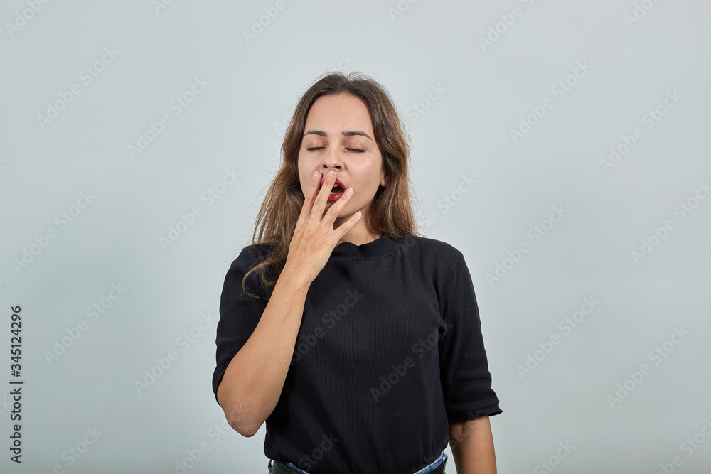 Cute Young Brunette Woman In Black T-Shirt, Blue Jeans With Belt On Gray Background, Tired Girl Yawns And Holds Her Hand To Her Mouth Cute Young Brunette Woman In Black T-Shirt, Blue Jeans With Belt On Gray Background, Tired Girl Yawns And Holds Her Hand To Her Mouth