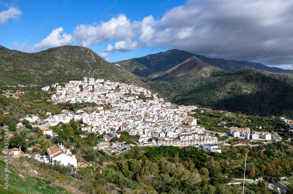 A Spanish village with white houses and red roofs is located on a mountainside against a sunny sky with clouds.