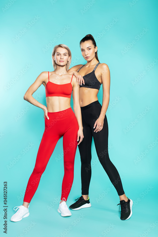 Fit and beautiful. Full length of two young and confident sporty girls in sportswear looking at camera while standing against blue background Fit and beautiful. Full length of two young and confident sporty girls in sportswear looking at camera while standing against blue background