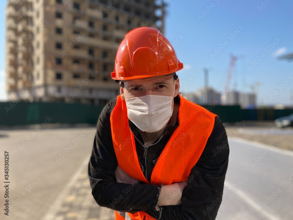 Portrait of male construction worker in medical mask and overalls on background of house under construction. Portrait of male construction worker in medical mask and overalls on background of house under construction.
