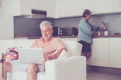 Stayhome and lockdown coronavirus old people concept - senior caucasian couple inside house in the kitchen cooking and use modern connected laptop computer - real life together